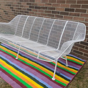 May include: A white metal bench featuring a grid-patterned wire mesh construction. The bench has a curved backrest, integrated armrests, and four thin, angled legs. The structure is composed of a continuous wire frame with a uniform white finish.