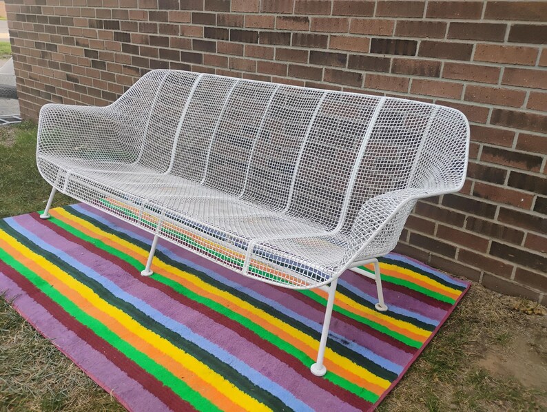 May include: A white metal bench featuring a grid-patterned wire mesh construction. The bench has a curved backrest, integrated armrests, and four thin, angled legs. The structure is composed of a continuous wire frame with a uniform white finish.