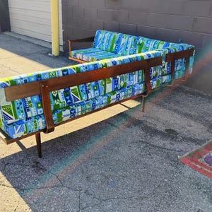 May include: A mid-century modern sectional sofa with a wooden frame and legs. The cushions are upholstered in a blue, green, and white geometric pattern. The sofa has a long, low profile and a built-in side table.