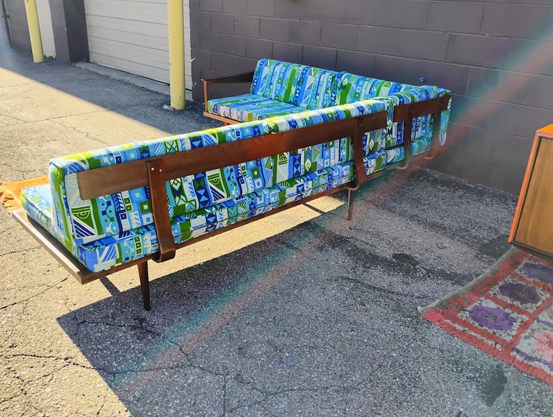 May include: A mid-century modern sectional sofa with a wooden frame and legs. The cushions are upholstered in a blue, green, and white geometric pattern. The sofa has a long, low profile and a built-in side table.