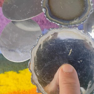 May include: A set of circular silver-toned coasters with scalloped edges and a reflective surface. The coasters feature a metallic finish with visible surface scratches and are arranged in a cluster, showing both the top and bottom sides.
