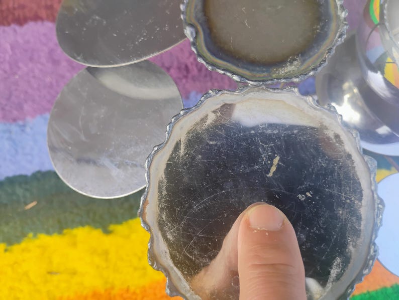 May include: A set of circular silver-toned coasters with scalloped edges and a reflective surface. The coasters feature a metallic finish with visible surface scratches and are arranged in a cluster, showing both the top and bottom sides.