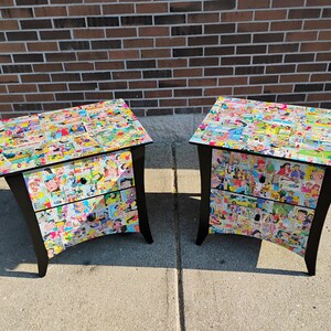May include: Two matching nightstands with black frames and comic strip-covered surfaces. Each nightstand has two drawers and a slanted design. The tops and drawer fronts are covered in colorful comic book images.