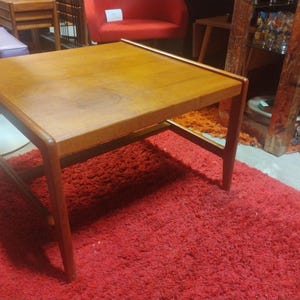 May include: A mid-century modern style coffee table with a wood top and a simple, sturdy frame. The table is sitting on a red shag rug.