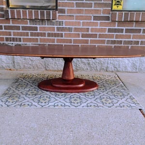 May include: A dark brown oval dining table with a pedestal base. The table is made of wood and sits on a patterned rug. The table is approximately 72 inches long.