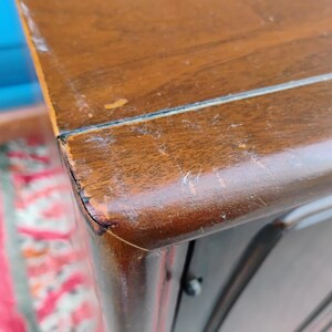 May include: A close-up view of the corner of a brown wooden cabinet with a smooth, polished finish. The surface shows visible signs of wear, including light scratches and minor paint loss along the sharp, angled edge of the furniture piece.