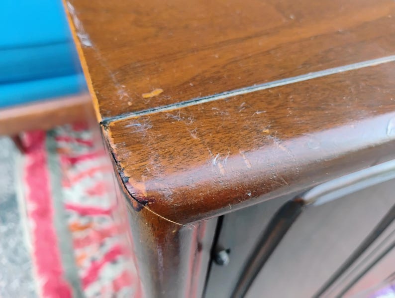 May include: A close-up view of the corner of a brown wooden cabinet with a smooth, polished finish. The surface shows visible signs of wear, including light scratches and minor paint loss along the sharp, angled edge of the furniture piece.