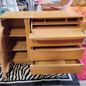 Op de afbeelding: Een licht houten kast met een neerlaatbaar bureau, drie lades en een kast met planken. De kast is open en de planken zijn leeg.