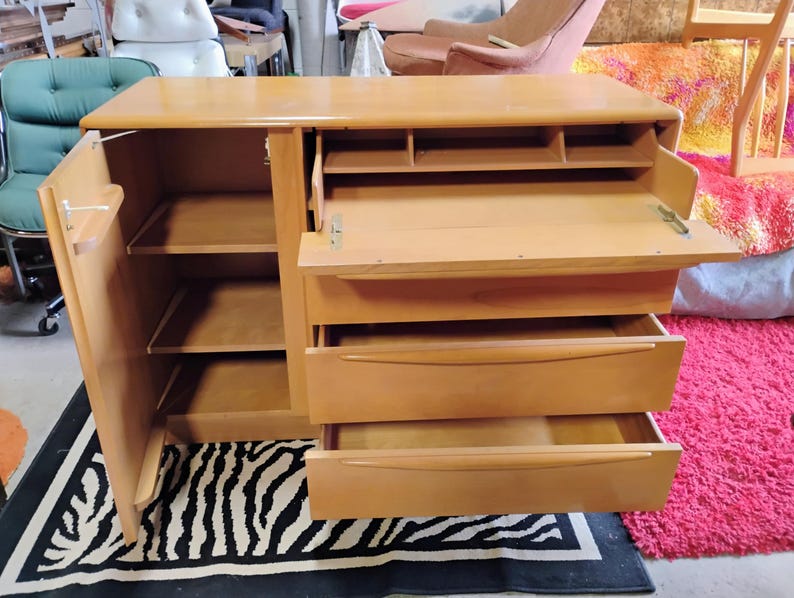 Op de afbeelding: Een licht houten kast met een neerlaatbaar bureau, drie lades en een kast met planken. De kast is open en de planken zijn leeg.