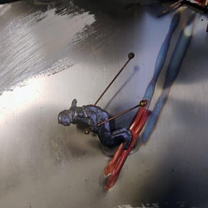 Pode incluir: Uma escultura em metal de um esquiador em posi&ccedil;&atilde;o de descida. O esquiador &eacute; azul escuro, com esquis cor de cobre. A escultura &eacute; montada sobre uma superf&iacute;cie de metal escovado. Os bast&otilde;es do esquiador s&atilde;o finas varetas de cobre com pequenas pontas em forma de bola.