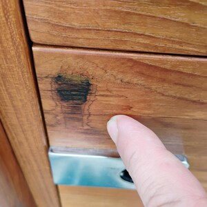 May include: A light brown wooden cabinet drawer featuring a prominent dark wood grain knot on the left side and a rectangular silver metal handle. The wood surface displays a smooth, polished finish with visible natural grain patterns throughout.