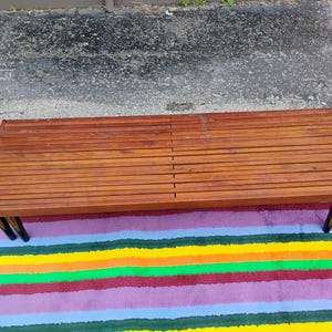 K&ouml;nnte beinhalten: Ein rechteckiger Couchtisch im Mid-Century-Modern-Stil mit einer Holzplatte mit Lamellen und schwarzen Beinen. Der Tisch steht auf einem bunten gestreiften Teppich. Das Holz hat einen warmen, r&ouml;tlich-braunen Farbton.
