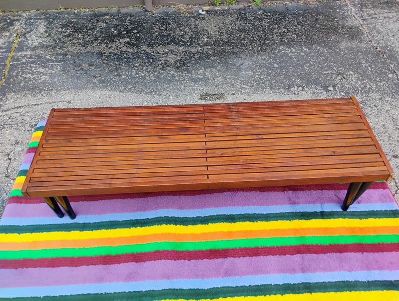K&ouml;nnte beinhalten: Ein rechteckiger Couchtisch im Mid-Century-Modern-Stil mit einer Holzplatte mit Lamellen und schwarzen Beinen. Der Tisch steht auf einem bunten gestreiften Teppich. Das Holz hat einen warmen, r&ouml;tlich-braunen Farbton.