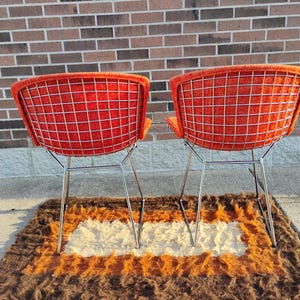 May include: Two mid-century modern chairs with bright orange seats and a grid-like wire back. The chairs have chrome legs and are set on a brown, orange, and white shag rug. The background is a brick wall.