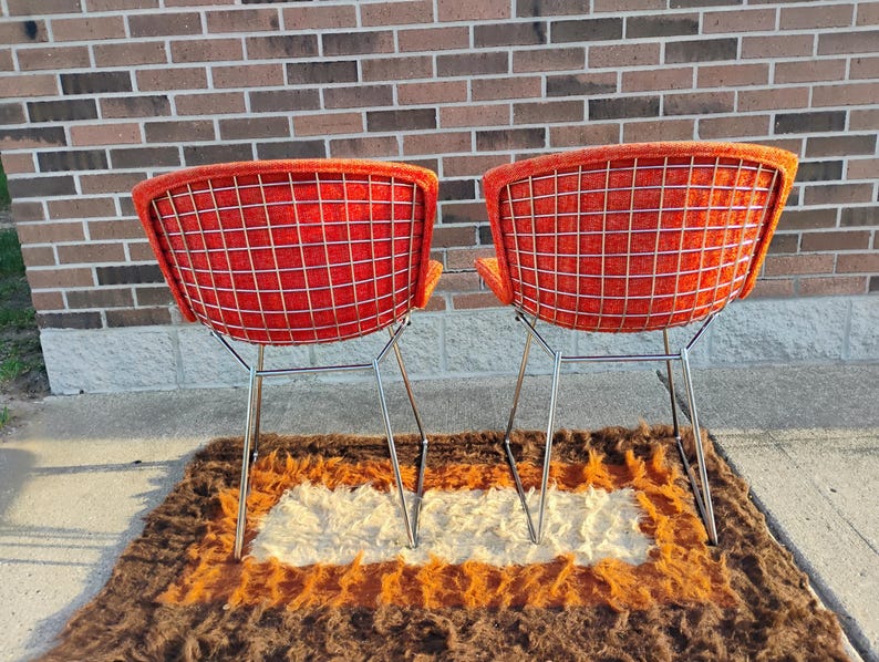 May include: Two mid-century modern chairs with bright orange seats and a grid-like wire back. The chairs have chrome legs and are set on a brown, orange, and white shag rug. The background is a brick wall.