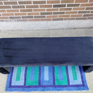 May include: A navy blue upholstered bench with a curved design. The bench is placed on a colorful rug with blue, green, and purple geometric patterns. The background features a brick wall.
