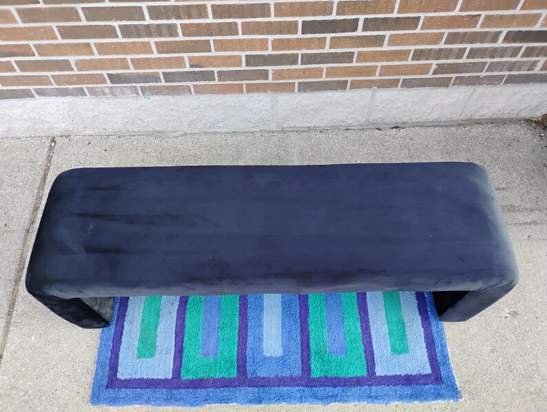 May include: A navy blue upholstered bench with a curved design. The bench is placed on a colorful rug with blue, green, and purple geometric patterns. The background features a brick wall.