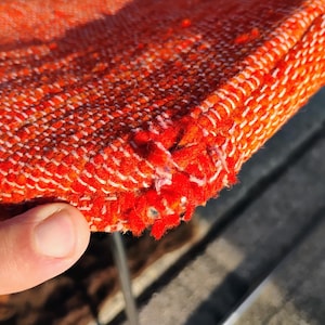 May include: Close-up of a section of orange woven fabric with white stitching along the edge. The fabric has a textured appearance and shows some wear and tear, with frayed edges.