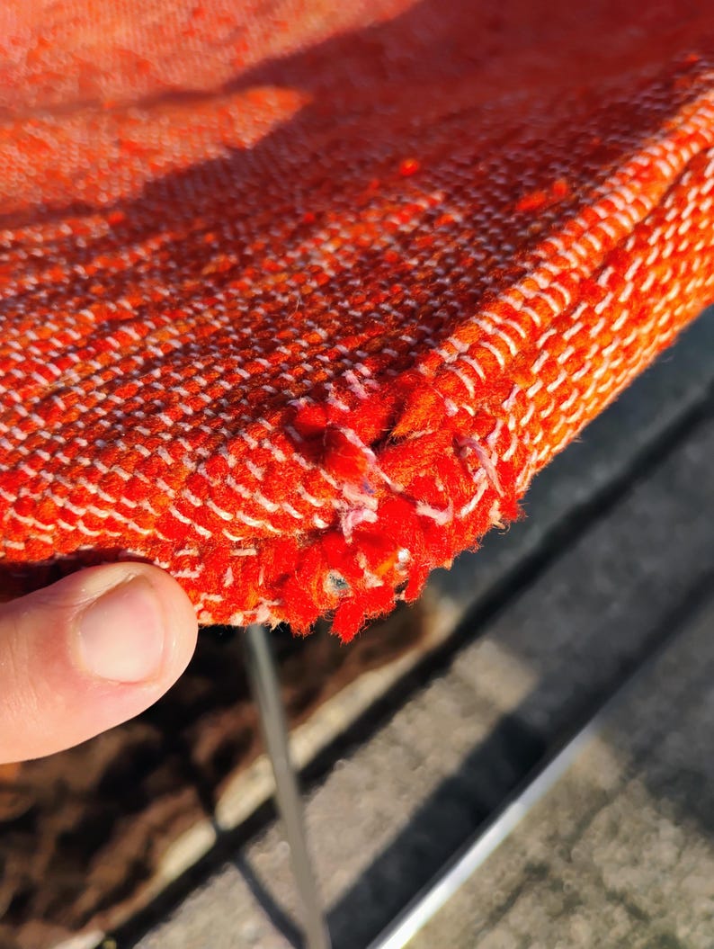 May include: Close-up of a section of orange woven fabric with white stitching along the edge. The fabric has a textured appearance and shows some wear and tear, with frayed edges.