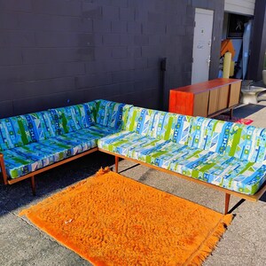 May include: A mid-century modern sectional sofa with a wooden frame and patterned cushions in blue, green, and white. An orange shag rug sits in front of the sofa. A wooden cabinet is in the background.