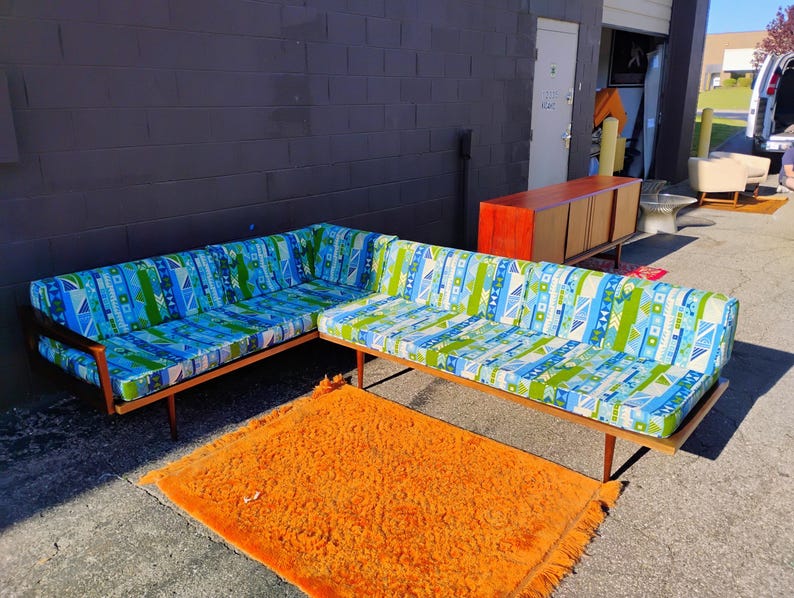 May include: A mid-century modern sectional sofa with a wooden frame and patterned cushions in blue, green, and white. An orange shag rug sits in front of the sofa. A wooden cabinet is in the background.