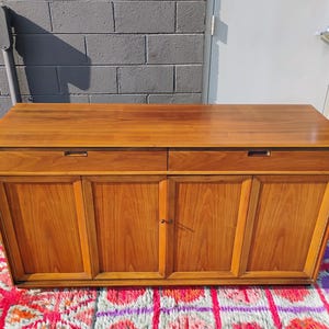 Op de afbeelding: Een rechthoekig houten dressoir met een glad bovenoppervlak, twee horizontale lades met verzonken handgrepen en vier onderste kastdeuren met verticale houtnerfpanelen in een middelbruine houtkleur.
