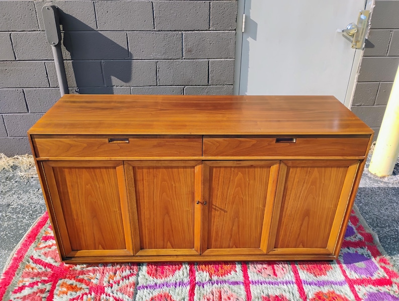 Op de afbeelding: Een rechthoekig houten dressoir met een glad bovenoppervlak, twee horizontale lades met verzonken handgrepen en vier onderste kastdeuren met verticale houtnerfpanelen in een middelbruine houtkleur.