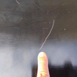 May include: A smooth, dark black surface featuring a series of visible, thin, light-colored scratches across the finish. The surface shows signs of wear with multiple fine lines and marks scattered across the dark, reflective material.