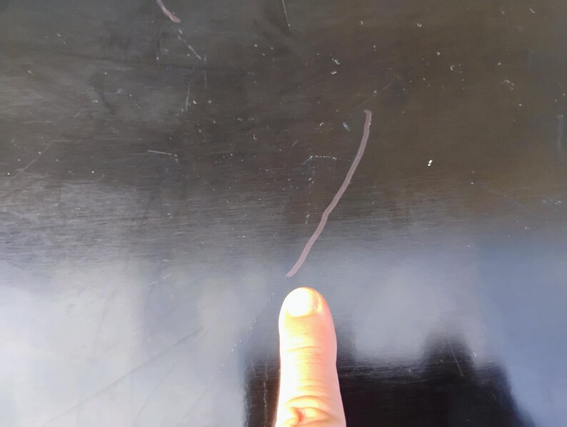 May include: A smooth, dark black surface featuring a series of visible, thin, light-colored scratches across the finish. The surface shows signs of wear with multiple fine lines and marks scattered across the dark, reflective material.