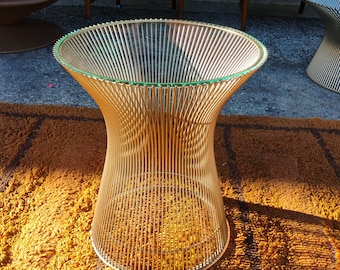 May include: A gold-colored wire side table with a clear glass top. The table has a unique, open design with vertical wires forming a flared shape. The table is sitting on a shag rug.