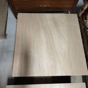 May include: A light-colored, unfinished wooden tabletop, likely plywood, with visible wood grain. The square table is positioned in a room with other furniture, including a wooden dresser and a patterned sofa.
