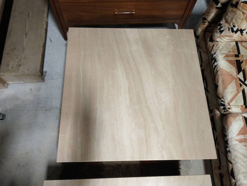 May include: A light-colored, unfinished wooden tabletop, likely plywood, with visible wood grain. The square table is positioned in a room with other furniture, including a wooden dresser and a patterned sofa.