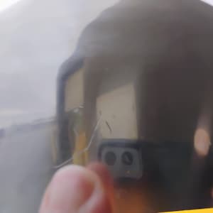 K&ouml;nnte beinhalten: Nahaufnahme einer reflektierenden, dunkelgrauen Oberfl&auml;che. Die Oberfl&auml;che ist leicht gekr&uuml;mmt, mit einigen Kratzern und Unvollkommenheiten. Eine verschwommene Reflexion einer Hand und eines Smartphones mit zwei Kameras ist sichtbar.