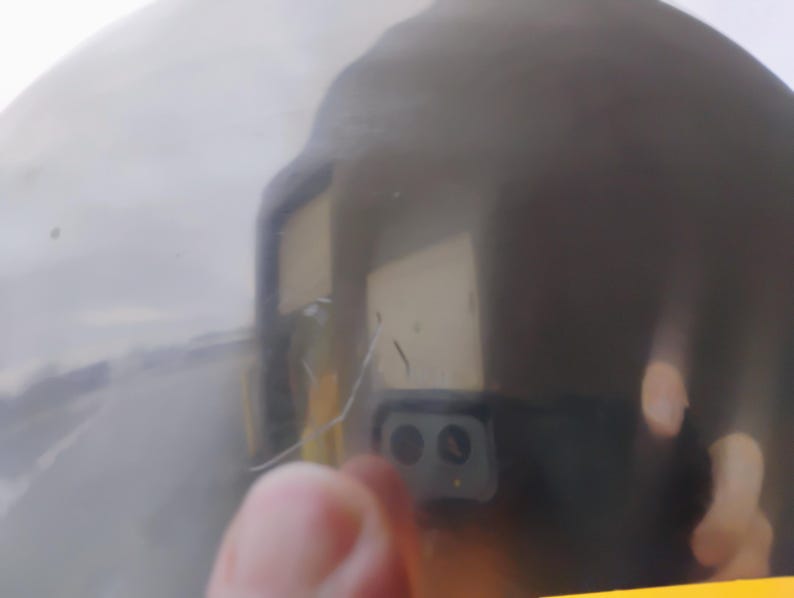 K&ouml;nnte beinhalten: Nahaufnahme einer reflektierenden, dunkelgrauen Oberfl&auml;che. Die Oberfl&auml;che ist leicht gekr&uuml;mmt, mit einigen Kratzern und Unvollkommenheiten. Eine verschwommene Reflexion einer Hand und eines Smartphones mit zwei Kameras ist sichtbar.