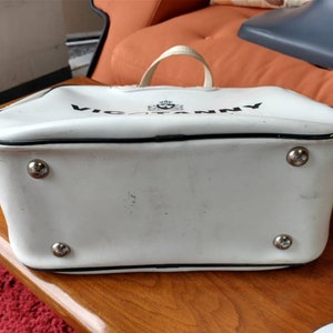 Pode incluir: Bolsa de m&atilde;o de vinil branco com acabamento preto e o texto "VICTORIANNY" impresso na frente. A bolsa tem quatro tachas de metal prateadas na parte inferior.