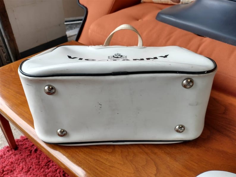 Pode incluir: Bolsa de m&atilde;o de vinil branco com acabamento preto e o texto "VICTORIANNY" impresso na frente. A bolsa tem quatro tachas de metal prateadas na parte inferior.