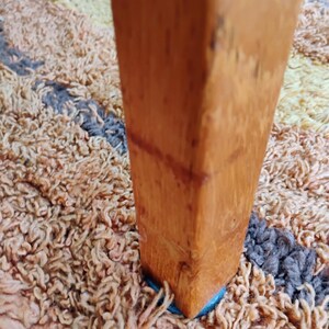 Op de afbeelding: Een verticaal, vierkant meubelpootje van hout met een mediumbruine afwerking en zichtbare houtnerf. De poot staat rechtop op een getextureerd, hoogpolig tapijt met een veelkleurig patroon van beige, bruine en donkergrijze vezels.