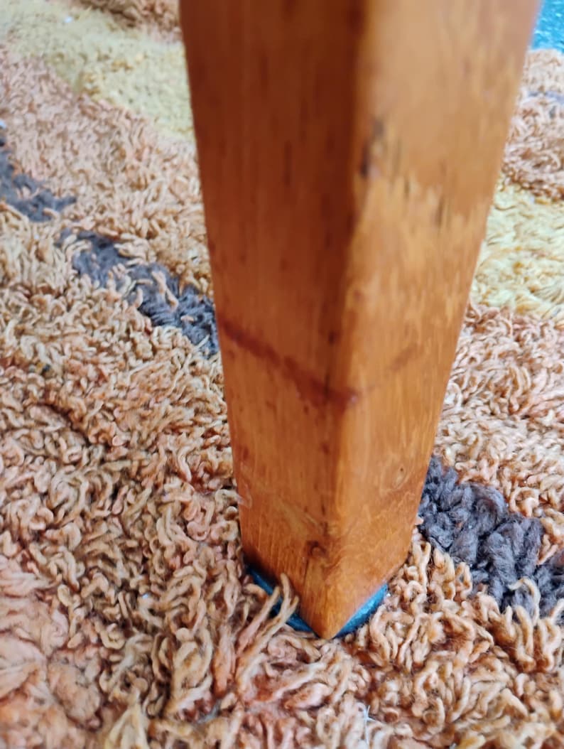 Op de afbeelding: Een verticaal, vierkant meubelpootje van hout met een mediumbruine afwerking en zichtbare houtnerf. De poot staat rechtop op een getextureerd, hoogpolig tapijt met een veelkleurig patroon van beige, bruine en donkergrijze vezels.