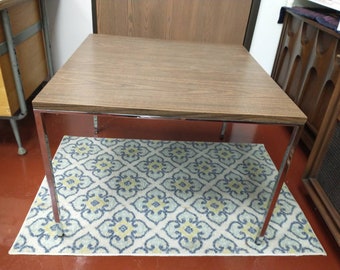 Könnte beinhalten: Ein quadratischer, brauner Holz-Couchtisch mit einem verchromten Gestell. Der Tisch steht auf einem gemusterten Teppich in den Farben Blau, Weiß und Gelb.