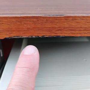 May include: A wooden desk surface with a visible horizontal scratch along the front edge, positioned above a light gray sliding drawer mechanism with metal side rails and a visible gap between the wood and the drawer component.