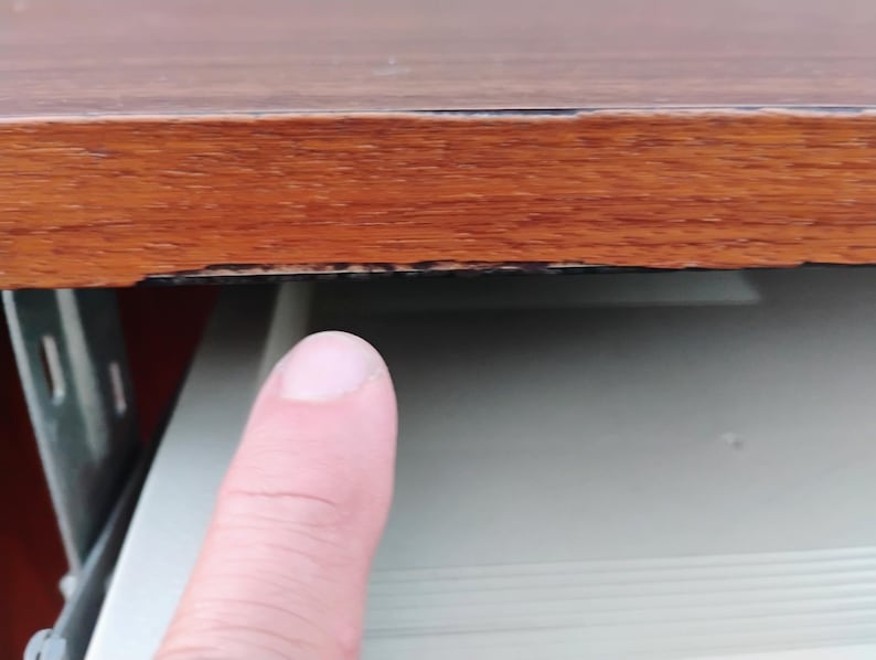 May include: A wooden desk surface with a visible horizontal scratch along the front edge, positioned above a light gray sliding drawer mechanism with metal side rails and a visible gap between the wood and the drawer component.