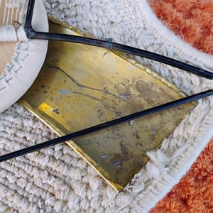May include: Close-up of a chair with a cream-coloured, stitched seat and a brass-coloured metal back. The chair's legs are black and the chair is resting on a textured, off-white rug with an orange border.