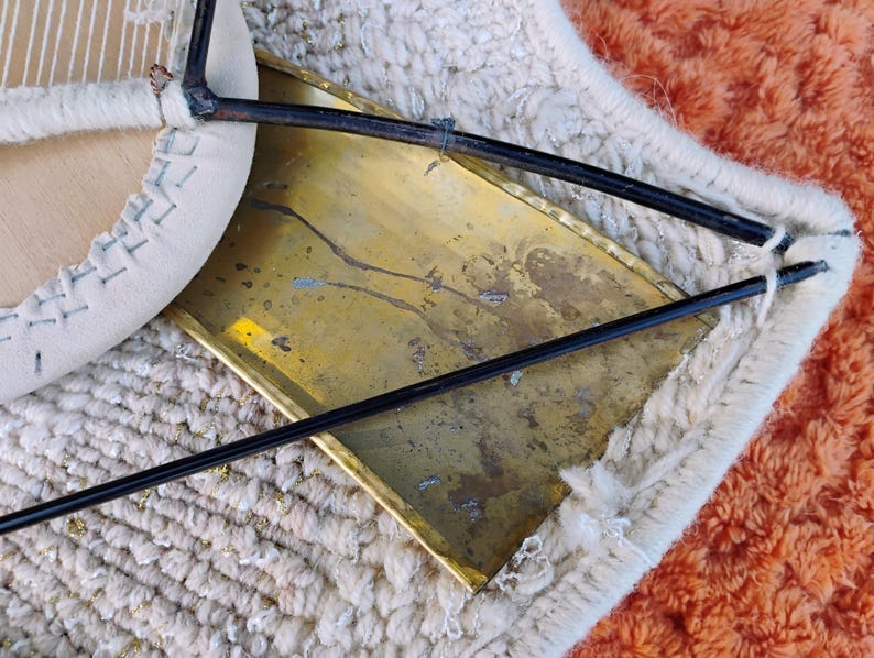 May include: Close-up of a chair with a cream-coloured, stitched seat and a brass-coloured metal back. The chair's legs are black and the chair is resting on a textured, off-white rug with an orange border.