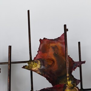 May include: Abstract metal wall sculpture with a geometric design. The sculpture features a grid of metal rods with irregular, colorful metal pieces attached. The pieces are painted in shades of red, orange, yellow, and brown.