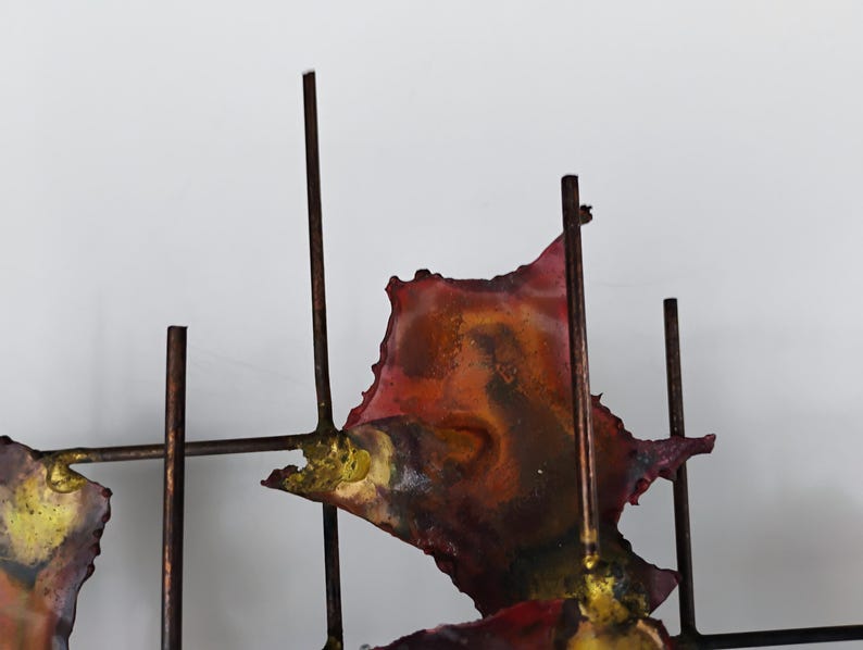 May include: Abstract metal wall sculpture with a geometric design. The sculpture features a grid of metal rods with irregular, colorful metal pieces attached. The pieces are painted in shades of red, orange, yellow, and brown.