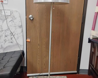 Könnte beinhalten: Eine Stehlampe mit gebürstetem Silber-Finish, bestehend aus einer hohen, schlanken Mittelsäule, einem runden Fuß und einem großen, kuppelförmigen Lampenschirm. Die Lampe verfügt über einen kleinen Zugschalter unter dem Schirm.