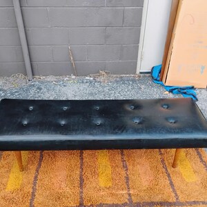May include: A rectangular black bench featuring a tufted seat with eight buttons arranged in two rows. The bench is supported by four tapered wooden legs that extend outward from the base, providing a stable foundation for the long, flat seat.