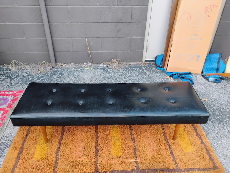 May include: A rectangular black bench featuring a tufted seat with eight buttons arranged in two rows. The bench is supported by four tapered wooden legs that extend outward from the base, providing a stable foundation for the long, flat seat.