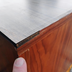 May include: A wooden cabinet featuring a light-colored, smooth top surface with a visible wood grain pattern. The side panels are constructed from a darker, reddish-brown wood veneer. A small gap is visible where the top meets the side panel.