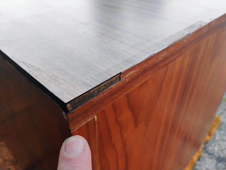 May include: A wooden cabinet featuring a light-colored, smooth top surface with a visible wood grain pattern. The side panels are constructed from a darker, reddish-brown wood veneer. A small gap is visible where the top meets the side panel.